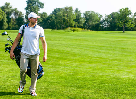 young male golfer carries a bag of golf clubs. Slim man in white T-shirt , golf shoes, white cap and beige pants is going in the green Turf Grass. Copy Space. Full-length. High quality photoの写真素材