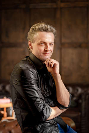 Charming Caucasian Man with Blue Eyes in a Black Shirt with Roll Sleeves Stands Half-Turn Looks at the Camera. Close-up Portrait. Wood Background. High quality photoの写真素材