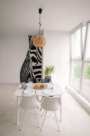 Interior Design of a Light Dining Room in a Minimalist Style with a Square Wooden Server White Table, Black and White Chairs, with Wicker Decorations on the Chandelier and on the Table, with a Large Window and a Huge Zebra Picture on the Wall. High quality photoの写真素材