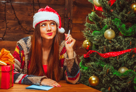 Charming Woman in a Red Hat with White Fur Sits at a Table near a Decorated Christmas Tree and Thinks what to Write to Santa. Close-up. High quality photoの写真素材