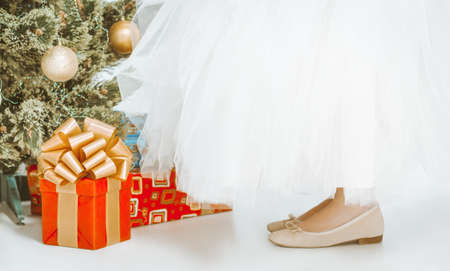 girlish legs in beige shoes and a long white tutu skirt stand on the floor near the Christmas tree. There are many gifts under the fir tree, square red gift box with gold ribbon and Big Loopy Bow. Close-up. High quality photoの写真素材