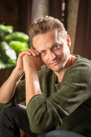 Charming Young Man with Blond Hair and Blue Eyes in Casual Clothes Looks Romantically at the Camera. Caucasian Male Model in Khaki Sweater Gazing at Camera. Close-up Portrait. High quality photoの写真素材
