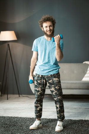 Smiling Young Man in Home Clothes does His Daily Workouts With Dumbbells at Home. Nice Man Pumps His Arm Muscles. High quality photoの写真素材