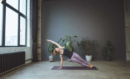 Lonely Woman in Sportswear Doing Side Plank in a Spacious Yoga Studio. Sporty Fit Woman Practices Hatha Yoga. Full Length. Gray Background. High quality photoの写真素材