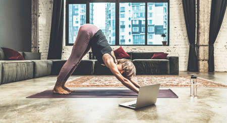 Nice Woman Practicing Yoga, Doing Down Dog Exercise, Watching Online Yoga Lessons by her Laptop, Yoga at Home. Yoga Time, Woman Relaxing in her Apartment in Megapolis City. Multi-storey Residential Building Backgroundの写真素材