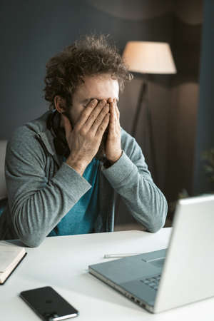 Tired Man Hiding his Eyes with Hands. He has Pain in his Eyes Because of Long-term Work at the Computer. Work Desk, Open Laptop, Mobile Phone. Close-up. High quality photoの写真素材