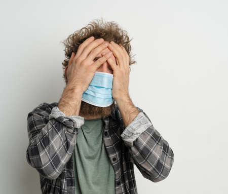 Caucasian Man in a Medical Mask has a Headache, Symptoms of Illness, Feeling Unwell, Malaise. White background. Close-up. Copy Space. High quality photoの写真素材