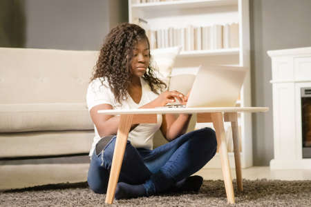Pretty Young Woman With Folded Legs Sitting on the Carpet and Typing on her Computer. Young Woman Looks Through the List of Films on Her Laptop. Close-up. Home Background . High quality photoの写真素材