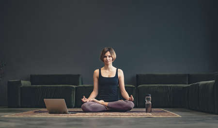 Young healthy woman sitting on the carpet with laptop and bottle of water in sportswear performing...