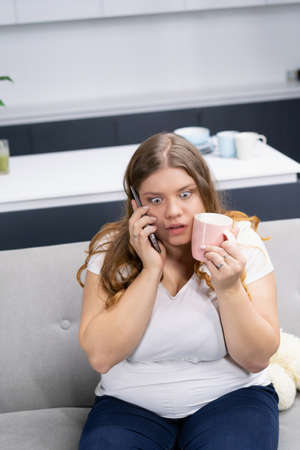 Scared or stressed overweight young girl talking on the phone holding pink cup pressed to her face. Fat or plus size girl talking on the phone sitting on the couch at home with a cup in hand.の写真素材