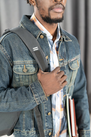 No face young african-american man with beard holding books and backpack strap wearing denim jacket isolated on grey. Handsome young student man on the way to school. School concept. Studies concept.の写真素材