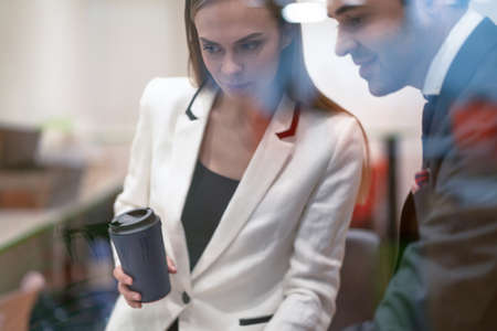 Two colleagues discuss project updates while having a coffee break. Young business people in the suits with coffee in hands looking down standing behind the glass.の写真素材