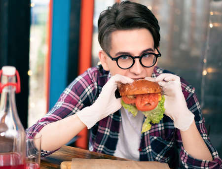 Teenager boy eats falling apart burger sitting at restaurant terrace. Handsome young man in glasses wearing rubber glows holding hamburger. Outdoor indoor street food restaurant. Street food concept.の写真素材