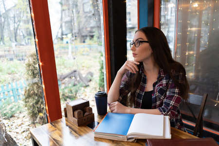 Charming girl study at university cafe wearing glasses and open books, notes in front of her. Young student girl in casual looking dreamed outside the cafe window drinking coffee from disposable cup.の写真素材