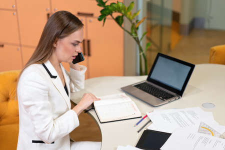 Business woman in white suit working in the office looking at diary or notebook with laptop and lots paper on the table. Bank manager looking at her schedule. HR manager updating her interview list.の写真素材