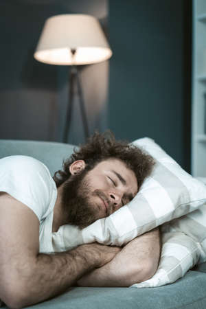 Handsome bearded young man sleeping on the sofa hugging a pillow after a long hours working late nigh. Sick young man sleeping on the couch after taking medications. distance work concept.の写真素材
