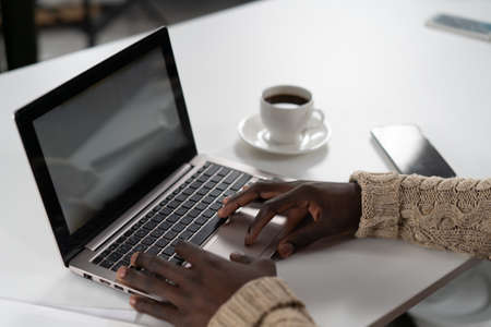 Young african freelancer man working on laptop while enjoying cup of coffee with smartphone next to him. African student study using laptop while coffee break at campus. Close up hands with laptop.の写真素材