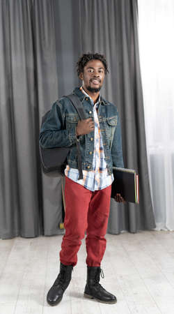 Trendy young african-american man with beard holding books and backpack wearing denim jacket ready to go to studies. Handsome young student man on the way to school. School concept. Studies concept.の写真素材