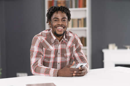 Smiling handsome African American man use smartphone checking social media or updating information. Portrait of young man holding smartphone sitting at the table. Social media concept.の写真素材