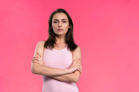 Suspicious young woman in a pink sleeveless t-shirt folded her arms and looks at the camera. Doubt,...