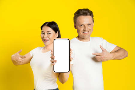 Caucasian man and asian woman show thumbs up and pointing finger excited holding smartphone with...