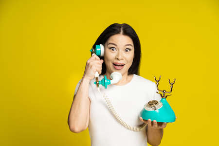 Funny face expression asian woman using vintage, retro telephone in hands wearing white t-shirt isolated on yellow background. Excited asian woman with retro phone. Communication concept.の写真素材