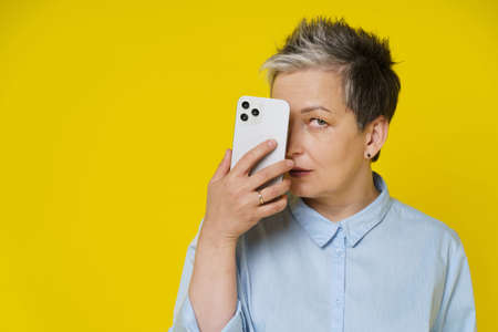 Grey haired mature woman hide face behind smartphone posting negative, toxic comment online, social media. Pretty woman with phone in blue shirt isolated on yellow background.の写真素材