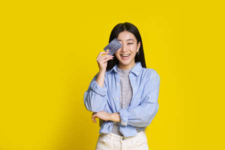 Happy excited young asian girl with credit, debit card. Asian girl in blue shirt holding mockup credit card isolated on yellow background. E-banking concept. E-commerce concept.の写真素材