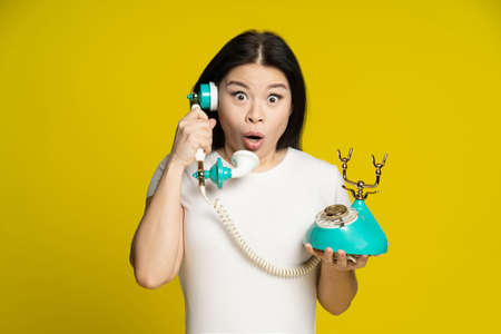 Asian woman talking using vintage, retro telephone in hands shock, excited, surprise face expression wearing white t-shirt isolated on yellow background. Communication concept.の写真素材