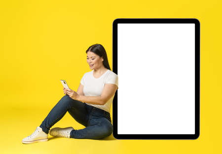 Asian woman leaned on huge, giant tablet pc sitting on floor with phone in hands in casual white screen, looking at phone mobile app advertisement isolated on yellow. Product placement.の写真素材