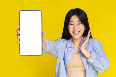 Amazed young asian girl showing or presenting mobile phone white white screen and excited smile on camera isolated on yellow background. Girl using smartphone. Selective focus on girl.の写真素材