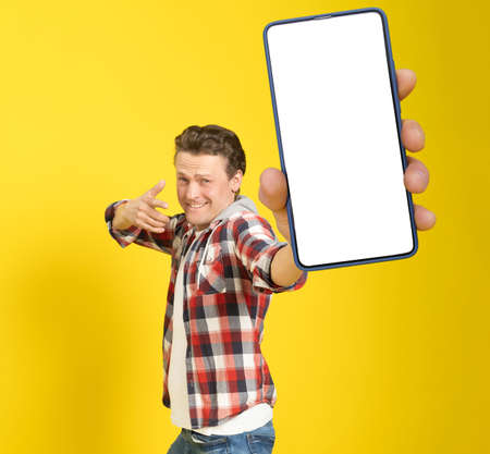 Happy to win handsome man pointing at smartphone with white empty screen, wearing red plaid shirt and jeans cellphone display mock up isolated on yellow background. Mobile app advertisement.の写真素材