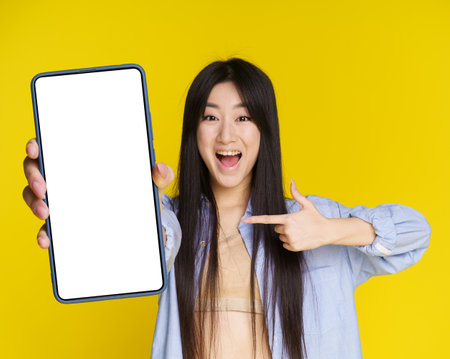 Asian girl excited win, lottery, casino, game, sale offer holding smartphone pointing on empty screen isolated on yellow background. Product placement mock up. Mobile application advertisement.の写真素材