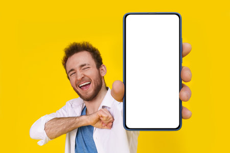 Yeah, chest hit happy excited young man with smartphone showing a white empty screen isolated over yellow game, bet, lottery win. Product placement. Mobile app advertisement.の写真素材
