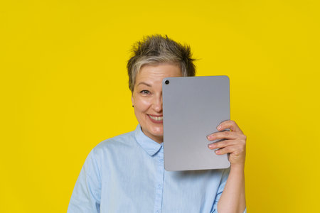 Grey haired mature woman with hide behind tablet pc working online or social media. Pretty woman in 50s wearing blue blouse isolated on yellow. Mature people and technologies.の写真素材