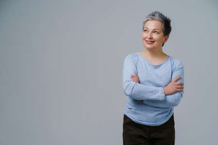 Mature grey hair woman posing with hands folded in blue blouse looking sideways up, copy space on left isolated on white background. Healthcare concept. Aged beauty concept. Copy space.の写真素材