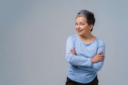 Challenging look positive emotions mature grey hair woman posing with hands folded looking at camera wearing blue blouse, isolated on white background. Healthcare, aged beauty concept. Copy space.の写真素材