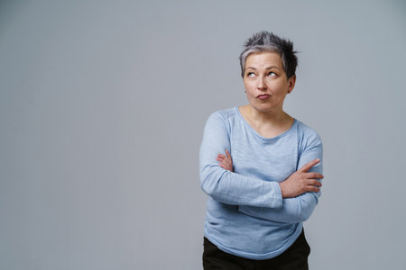 Pensive, thoughtful, doubt mature woman with grey hair in 50s posing with hands folded and copy space on left isolated on white background. Place for product placement. Aged beauty. Toned image.の写真素材