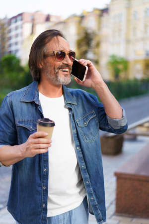 Excited mature man with grey beard talking on the phone while having coffee in paper disposable cup, work or travel time standing outdoors in urban city background wearing jeans shirt and sunglasses.の写真素材