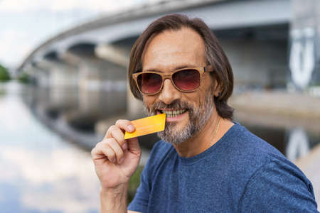 Handsome middle aged man standing outdoors at urban city background near the bridge over the river holding debit or credit card, shopping online or paying online banking. E-banking concept.の写真素材