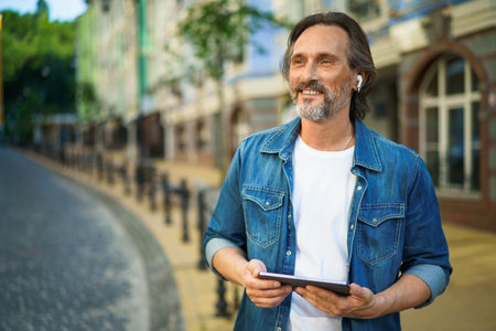 Handsome mature freelancer man with digital tablet in hands having a call while standing on urban streets. Mature man talking or listening music use wireless earphones while travel old town streets.の写真素材