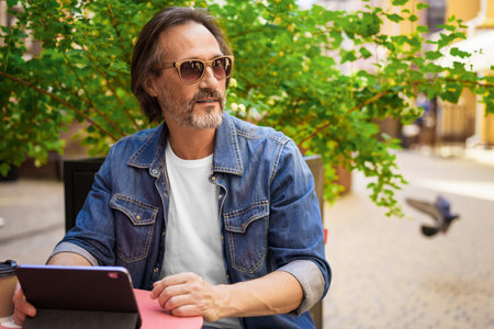 Middle aged man looking away sitting at cafe or street restaurant while working outdoors using digital tablet. Handsome mature freelancer man enjoying coffee outdoors while having group chat.の写真素材