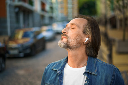 Enjoying life listening music with eyes closed standing outdoors old town street handsome mature grey bearded man with wireless earphones wearing casual outfit. Travel lifestyle concept.の写真素材