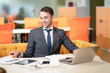 Young business man in suit, sitting at his desk, looking at documents. With laptop on the table, documents, papers, office supplies, a smartphone and tablet pc. Business concept.の写真素材