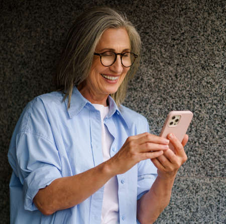 Charming mature grey hair woman talking on video call or reading text message standing outdoors...