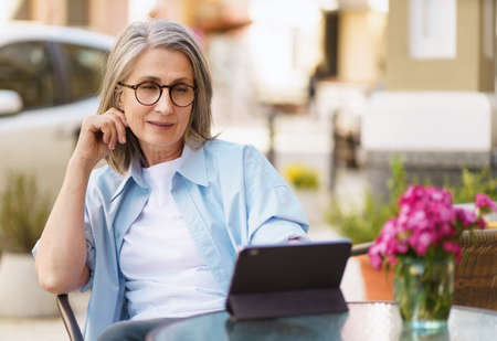 Charming european gray haired woman surf internet using digital tablet sitting at small street cafe. Mature silver hair woman working outdoors city cafe, wearing white tshirt and blue shirt.の写真素材
