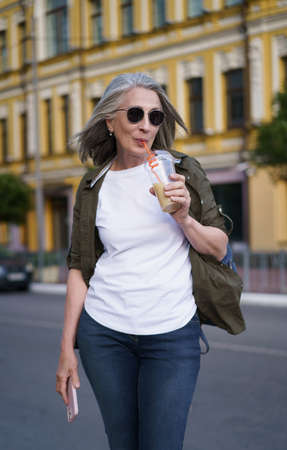 Senior european woman with gray hair happy enjoying free time traveling holding phone while having juice on the go using plastic cup in city background. Enjoying life mature woman.の写真素材