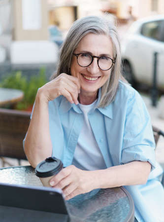 European gray haired mature woman drinks coffee while surf internet using digital tablet sitting at small street cafe. Mature woman working outdoors city cafe, wearing white tshirt and blue shirt.の写真素材