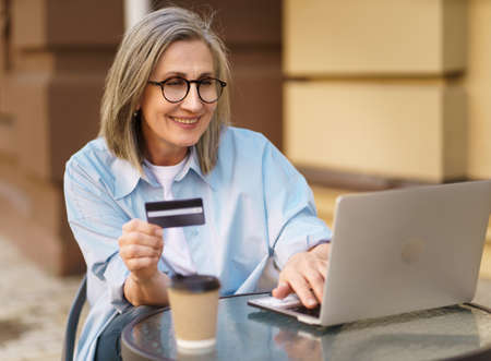 Charming mature european woman shopping or make online payment using laptop sitting outdoors street cafe terrace holding debit, credit card in hand. Mature woman happy vacation time traveling.の写真素材