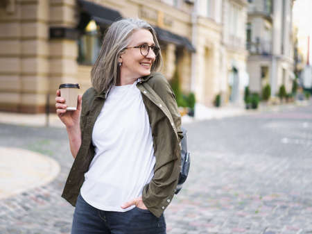 Businesswoman hold disposable cup with hot coffee. Mature woman spend free time traveling in european old town enjoying cup of tea, coffee. Grey haired woman in casual with backpack.の写真素材